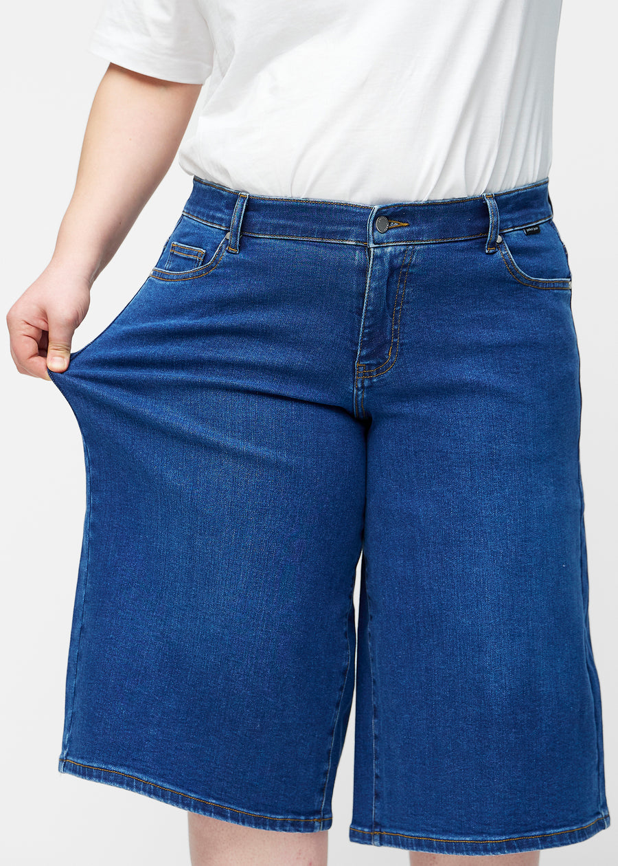 Plus-size model strækker de løse, mørkeblå denim capris i farven Oceans ved hoften for at vise materialets stretch.