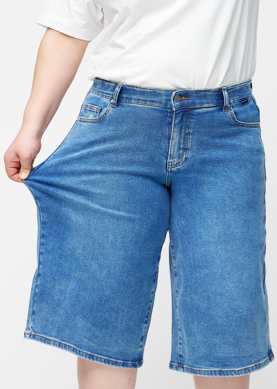 Plus-size model strækker de løse, klassisk blå denim capris i farven Rivers ved hoften for at vise materialets stretch.