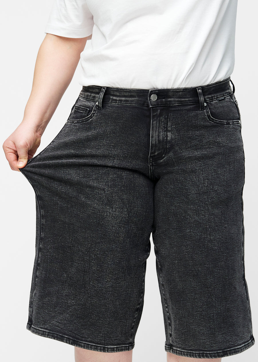 Plus-size model strækker de løse, mørkegrå denim capris i farven Shadows ved hoften for at vise materialets stretch.