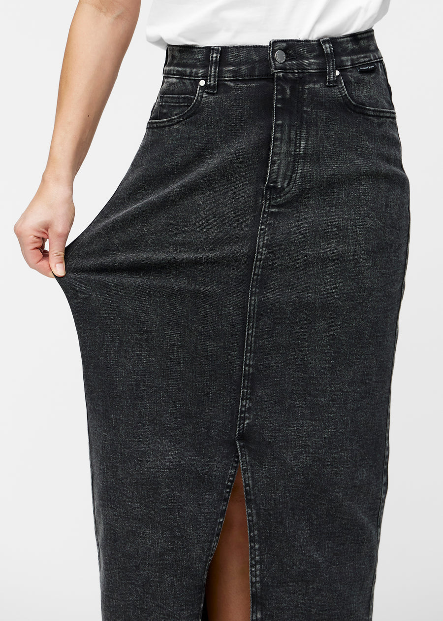 Model strækker den lange, mørkegrå denimnederdel i farven Shadows ved hoften for at vise materialets stretch.