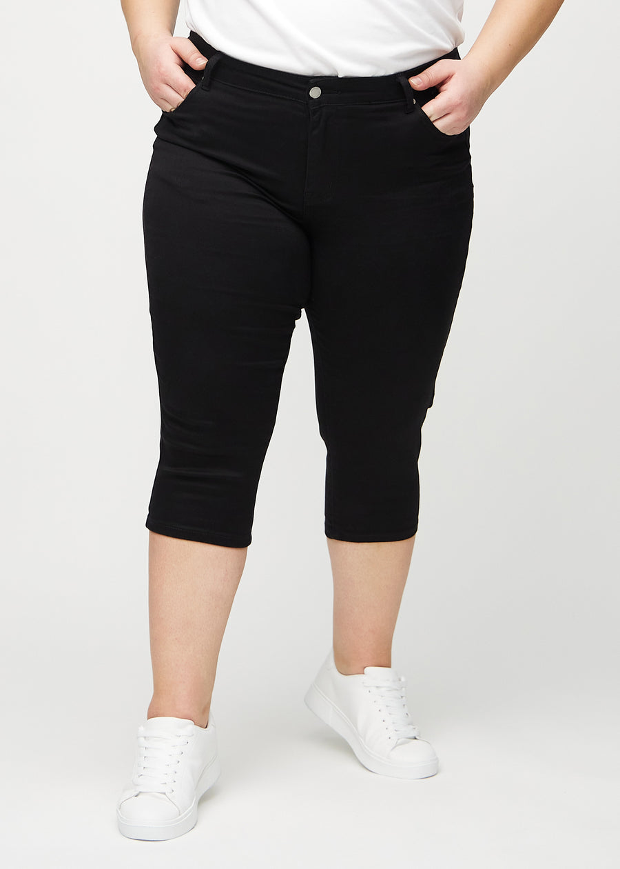 Black skinny capris, model name Ravens, which fit closely to the leg and end just below the knee on a plus-size model, seen from the front.
