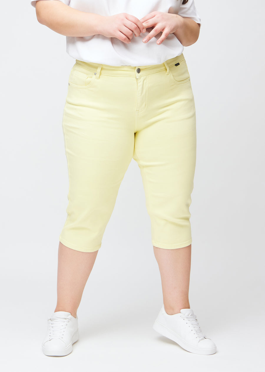 Slim capris for women in pastel light yellow jeans fabric, color Lemonades. Front view on a plus-size model, showing the fitted cut ending just below the knee.