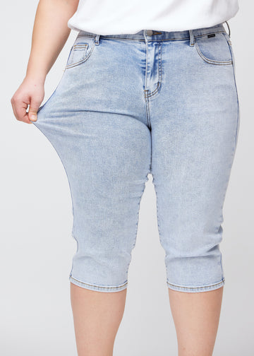 Slim capris for women in light blue denim, color Waves. A plus-size model pulls the fabric at the thigh to demonstrate stretch and flexibility.