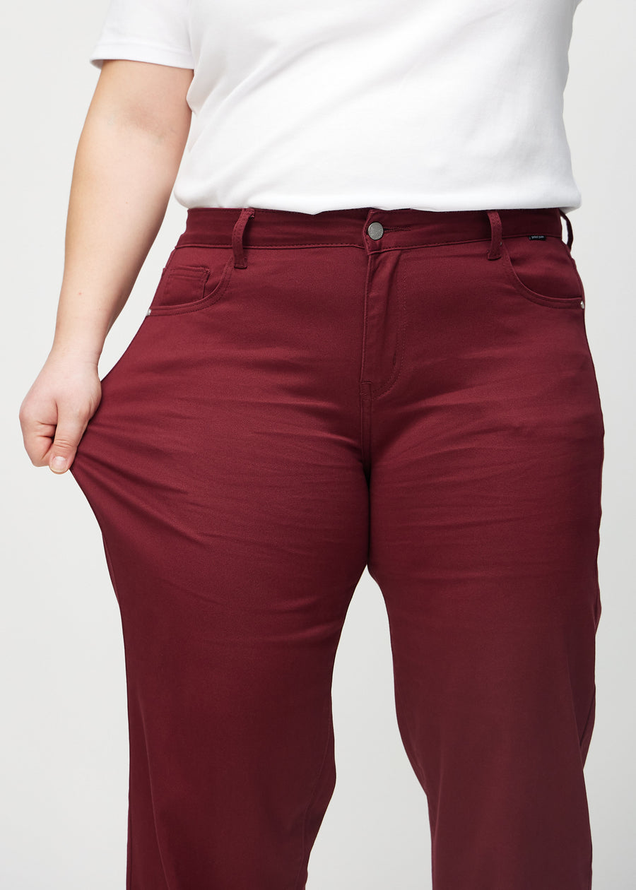 Loose jeans for women in dark red, color Santas. A plus-size model stretches the denim at the thigh to show the fabric&
