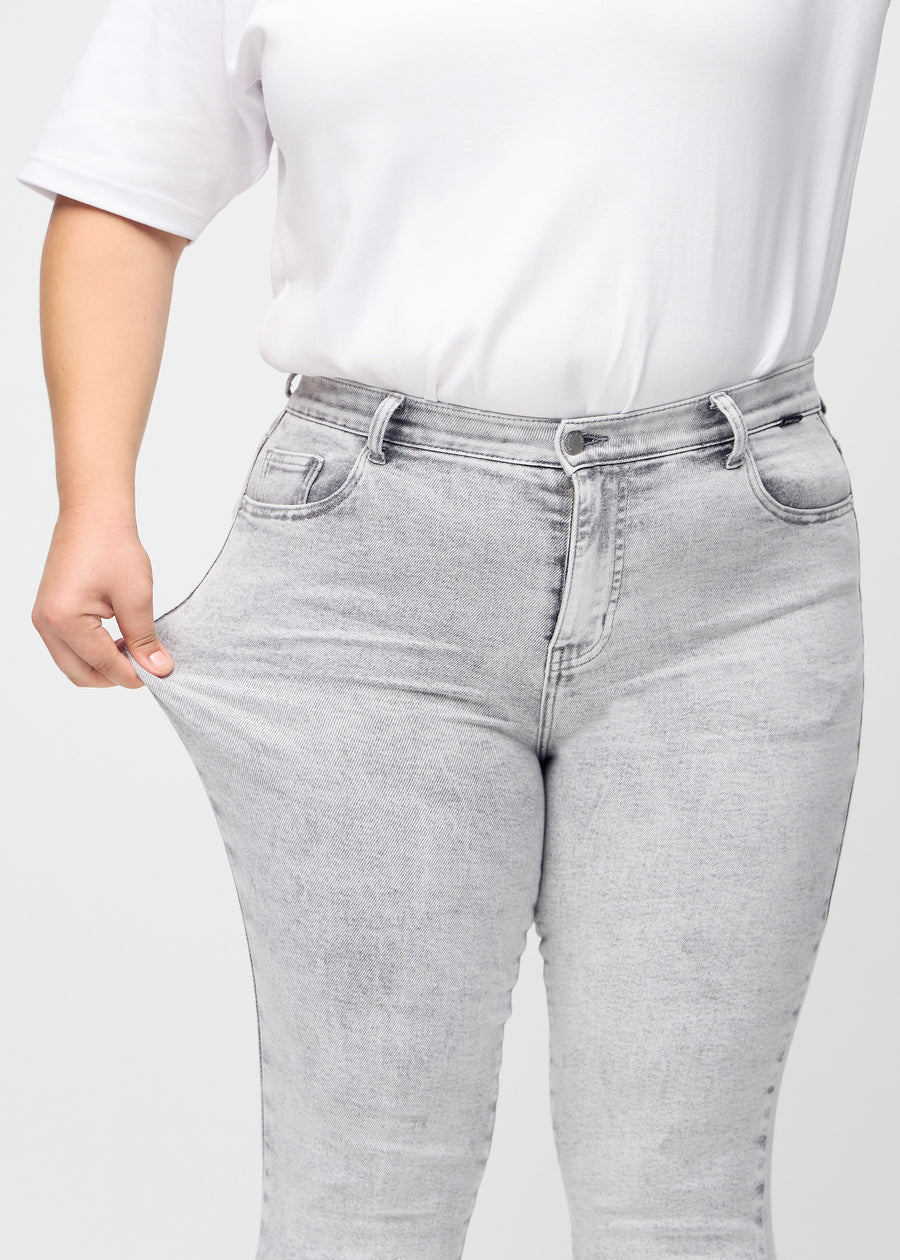 Skinny jeans for women in light grey, color Ashes. A plus-size model stretches the jeans at the thigh to show the fabric&