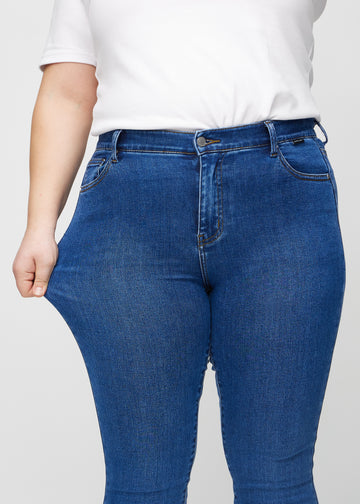 Skinny jeans for women in dark blue, color Oceans. A plus-size model stretches the jeans at the thigh to show the fabric&