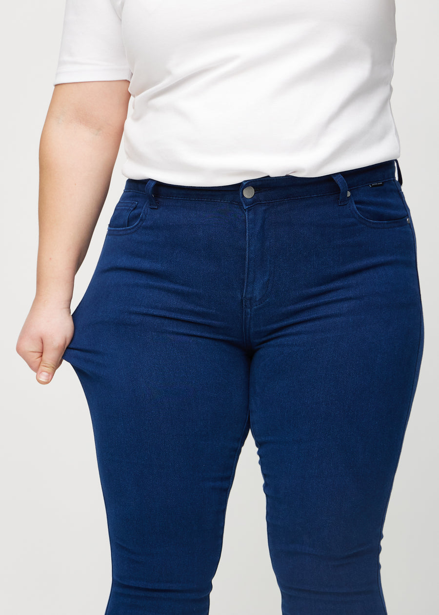 Skinny jeans for women in dark blue, color Royals. A plus-size model stretches the jeans at the thigh to show the fabric&
