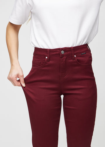 Slim jeans for women in dark red, color Santas. A model stretches the jeans at the thigh to show the fabric&