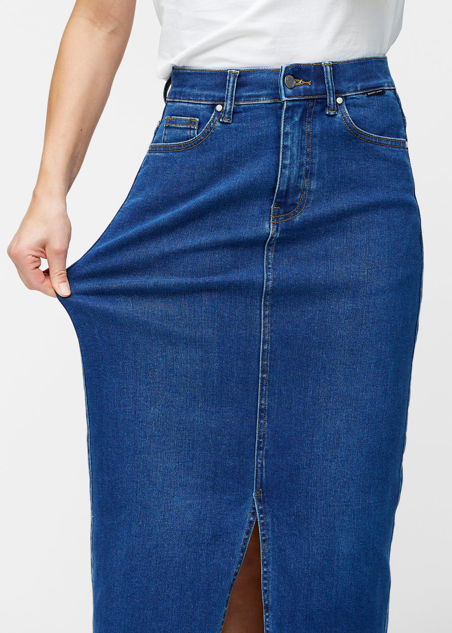 Model strækker den lange, mørkeblå denimnederdel i farven Oceans ved hoften for at vise materialets stretch.