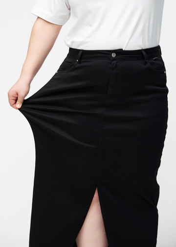 Plus-size model strækker den lange, sorte jeansnederdel i farven Ravens ved hoften for at vise materialets stretch.